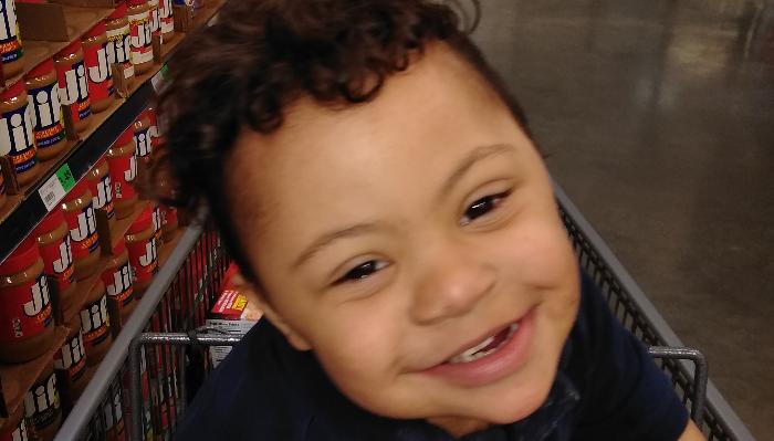 A smiling young boy with curly hair sitting in a shopping cart in a grocery store aisle.