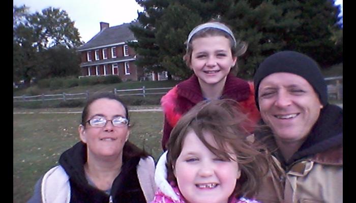 A happy family of four smiling together for an outdoor selfie in front of a brick house.