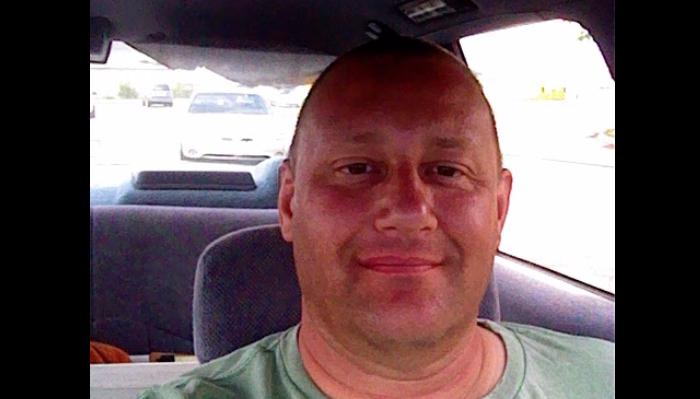 Close-up selfie of a smiling middle-aged man sitting inside a car wearing a green t-shirt.