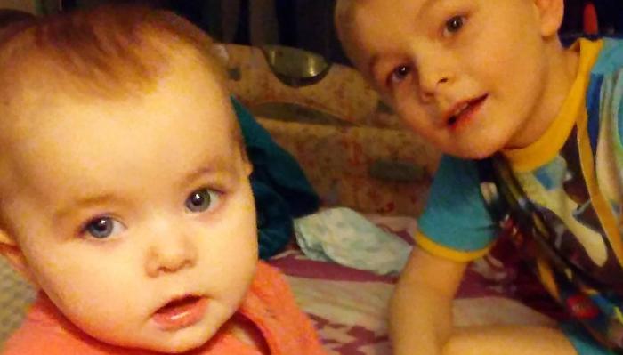 Close-up of a blue-eyed baby in an orange top sitting beside a young boy looking at the camera.