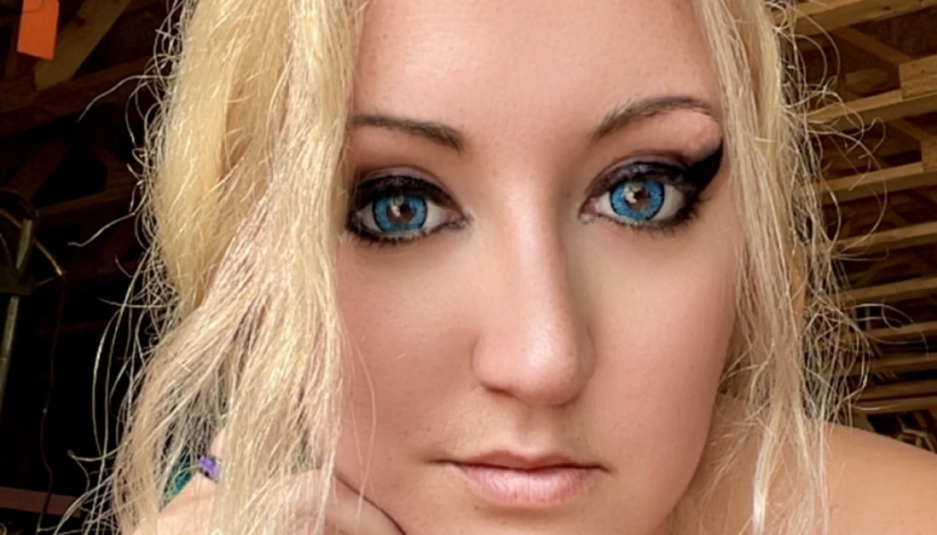 Close-up portrait of a blonde woman with striking bright blue eyes and dark eyeliner, looking at the camera.