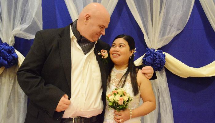 A smiling bride and groom gaze lovingly at each other, the bride holding a pink and white rose bouquet.