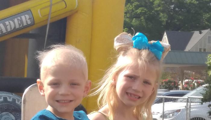 A smiling young boy and a girl with a large blue bow posing outdoors near a yellow bounce house.