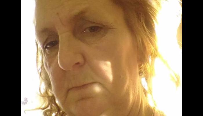 Close-up portrait of an older woman with reddish-blonde hair looking at the camera, backlit by bright sunlight.