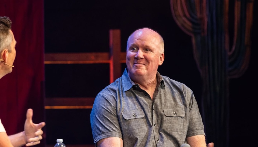 Smiling bald man wearing a grey short-sleeved shirt sitting on stage listening to an interviewer.