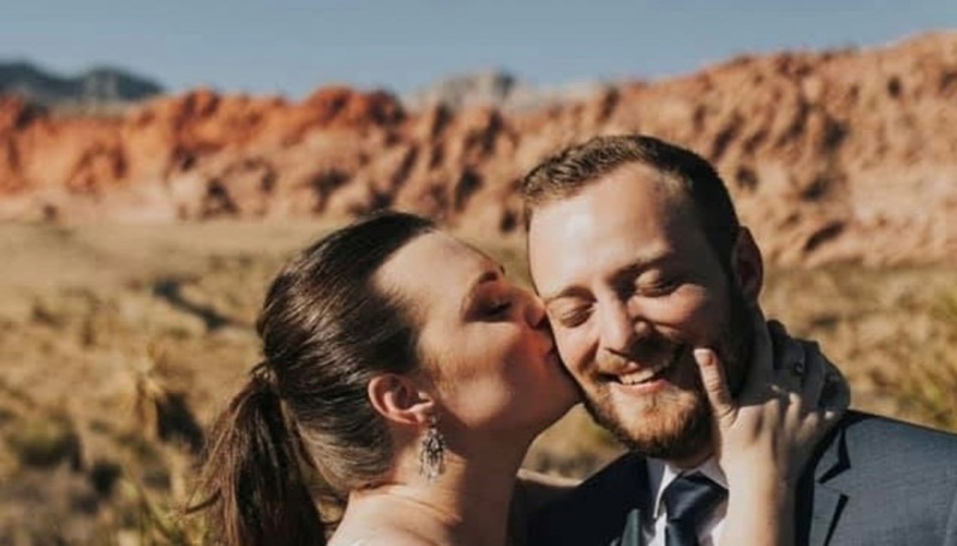 A bride kissing her smiling groom on the cheek with red rock mountains in the background.