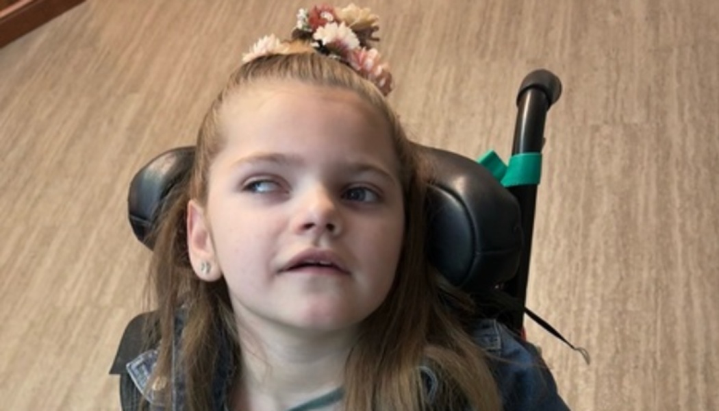 A young girl with a floral hairpiece sitting in a specialized wheelchair with head supports.