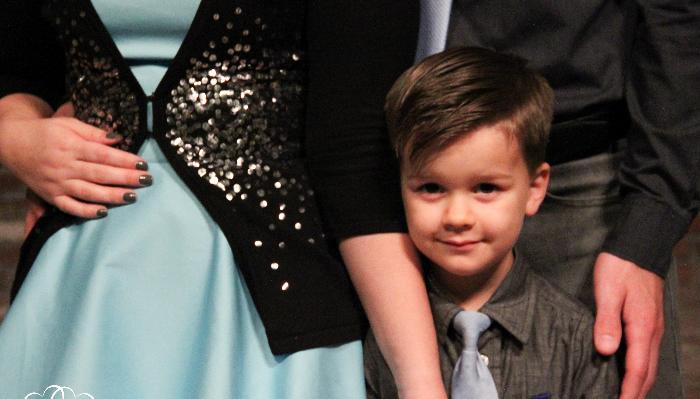 A young boy wearing a light blue tie stands smiling softly between his parents.