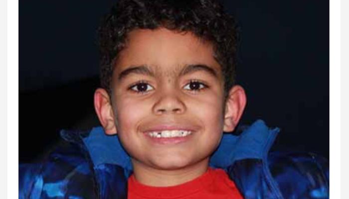 A smiling young boy with short curly dark hair wearing a blue jacket over a red t-shirt.