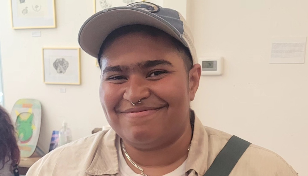 A smiling person wearing a baseball cap and nose ring stands in front of a white wall with framed art.