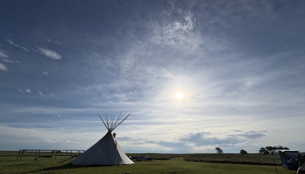 A traditional tipi stands in a vast grassy field beneath a bright sunny sky with wispy clouds.