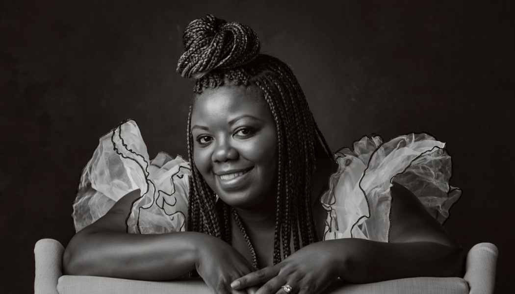 A smiling Black woman with braided hair in a top bun, wearing a ruffled outfit in a black-and-white portrait.