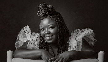 A smiling Black woman with braided hair in a top bun, wearing a ruffled outfit in a black-and-white portrait.