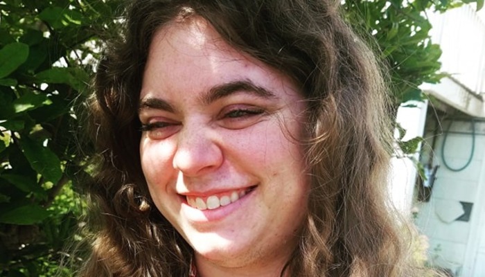 A smiling person with long curly brown hair outdoors in bright sunlight.