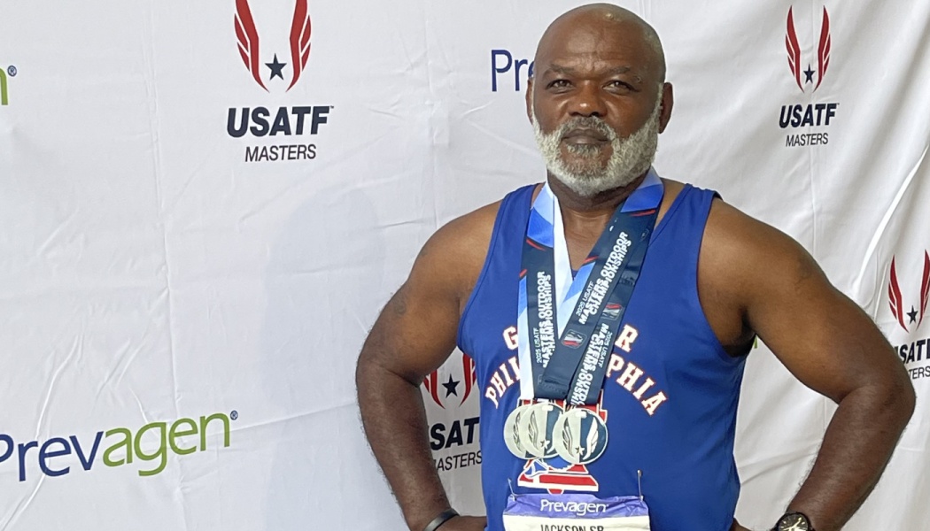 Senior male track athlete with a grey beard wearing three medals and a blue singlet in front of a USATF backdrop.