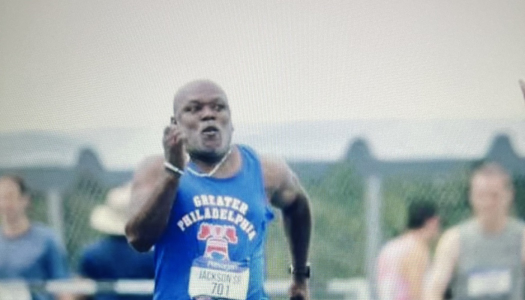 A male sprinter in a blue Greater Philadelphia singlet running with determination during a track race.