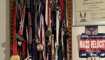Dozens of track and field award medals hanging on a rack next to a framed All-American certificate and race bib.