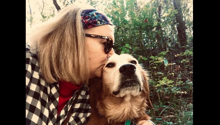 A woman wearing sunglasses and a headband lovingly kisses the top of her golden retriever's head outdoors.