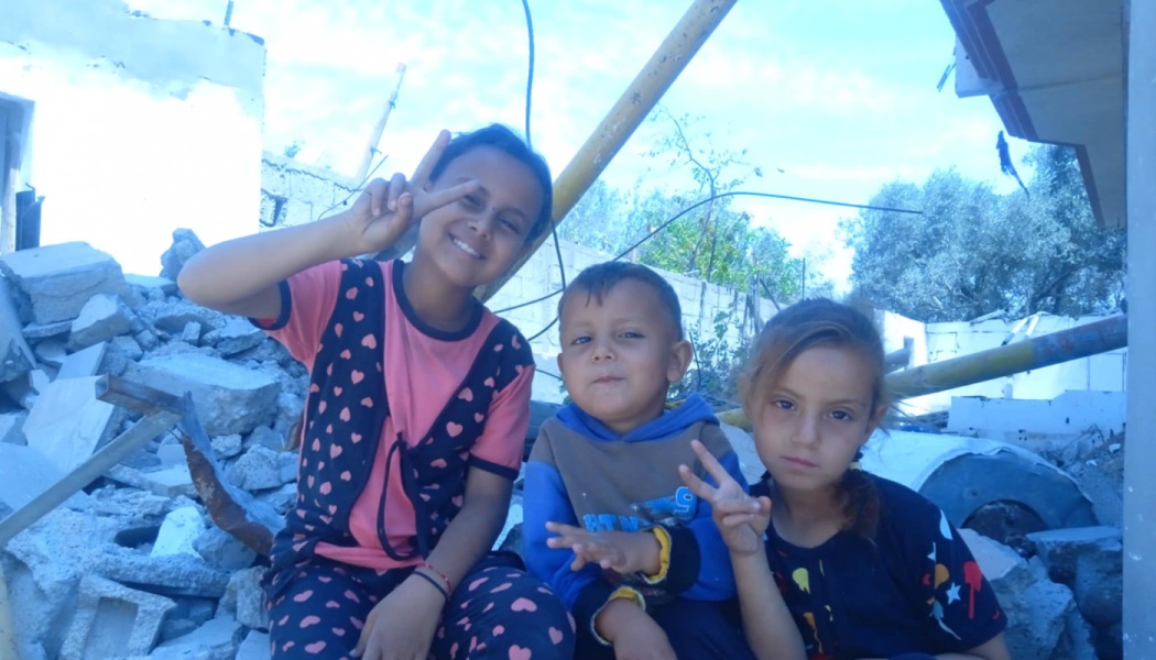 Three young children sit amidst destroyed building rubble, smiling and making peace signs.