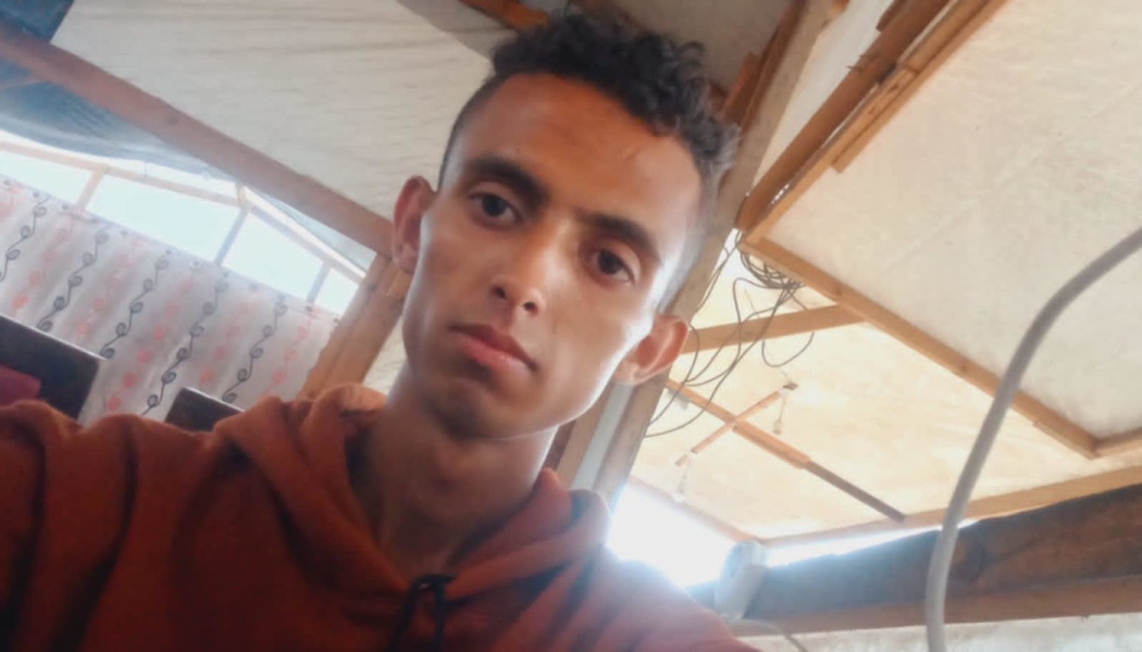 A young man with dark curly hair wearing a rust-brown hoodie looks at the camera inside a wooden structure.