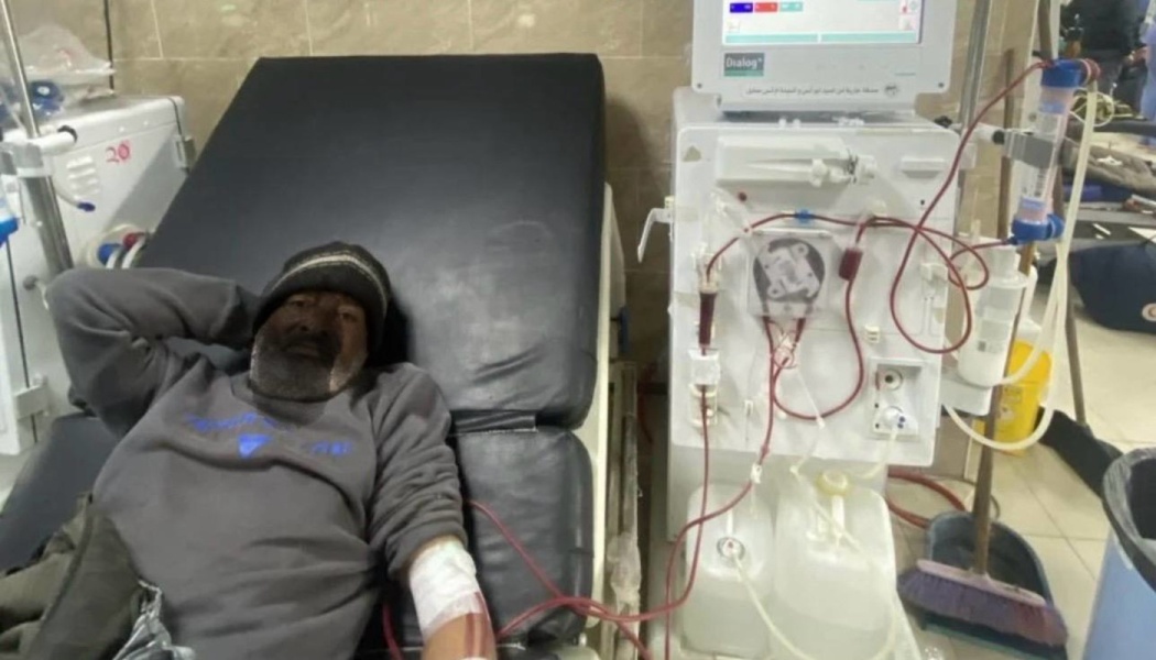 A bearded man in a beanie lies on a hospital bed connected to a dialysis machine.