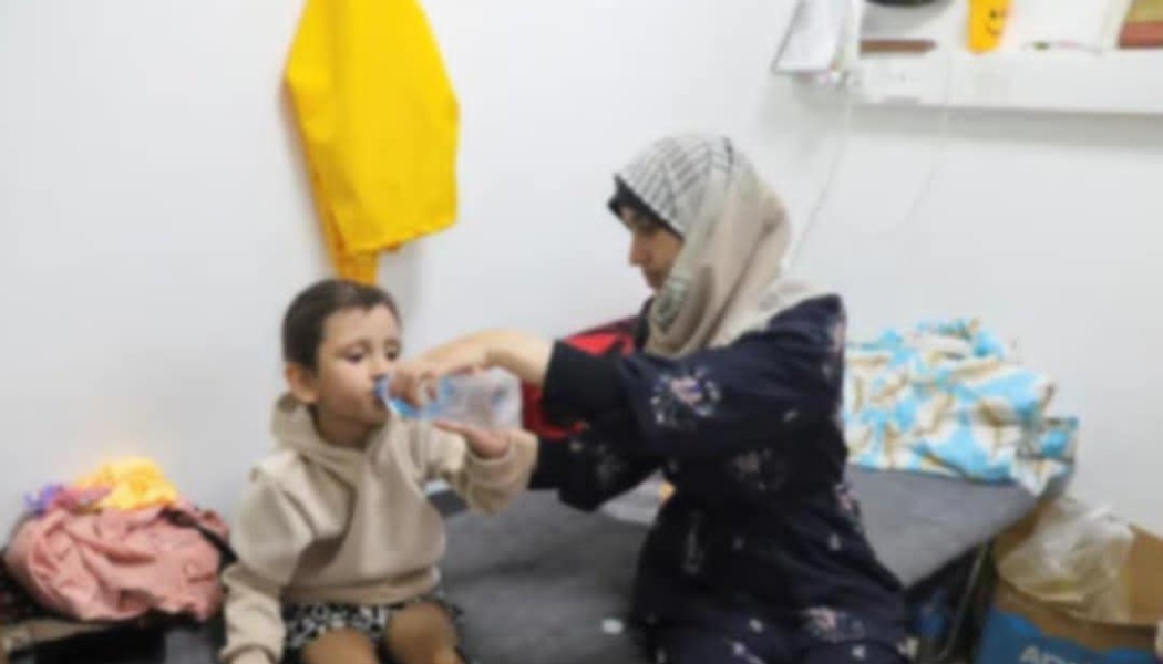 A woman wearing a hijab helps a young child drink water from a bottle inside a room.
