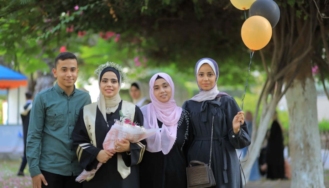 A smiling female graduate holding flowers stands outdoors with three family members, one holding balloons.