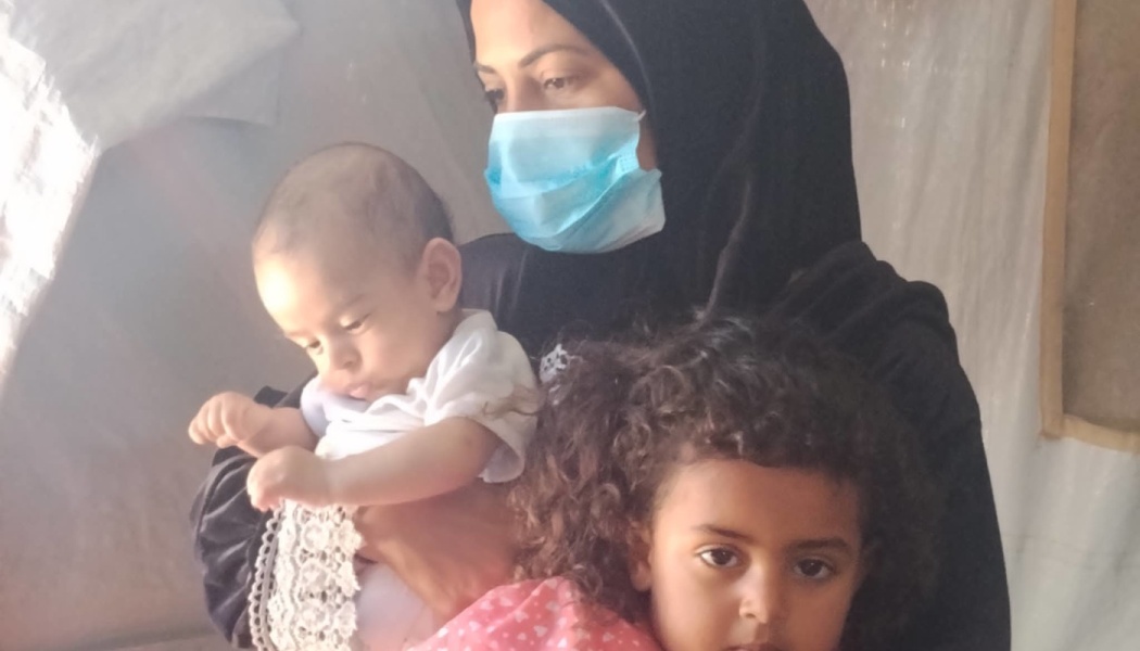 A woman wearing a black hijab and face mask holds a baby and a young girl with curly hair.