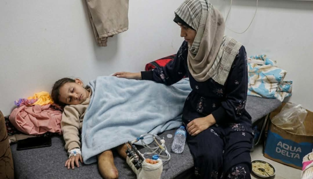 A mother in a hijab sits beside a young child lying on a cot, recovering from severe leg injuries.