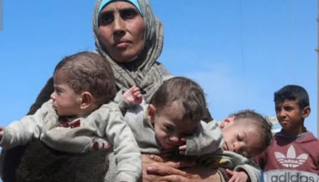 A woman in a headscarf holding three infants in her arms outdoors, with a young boy standing beside them.