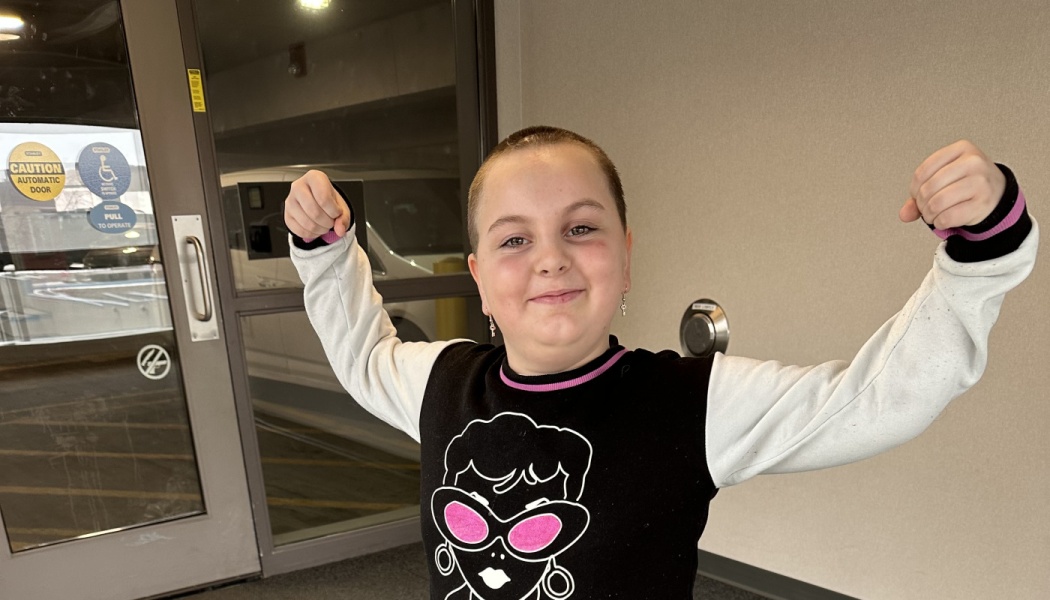 A smiling young child with a shaved head proudly flexes both arms in a strong pose.