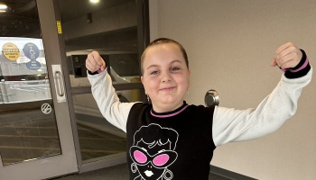 A smiling young child with a shaved head proudly flexes both arms in a strong pose.