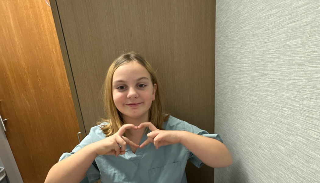Young girl in blue medical scrubs smiling and making a heart shape with her hands.
