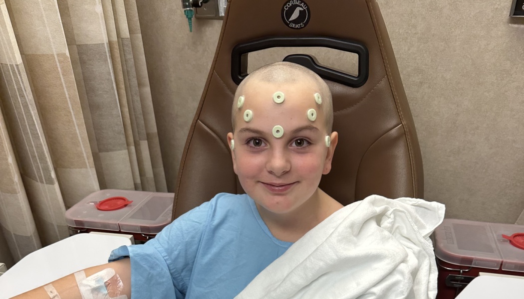 A smiling child with a shaved head sits in a medical chair with round sensors on their forehead during treatment.
