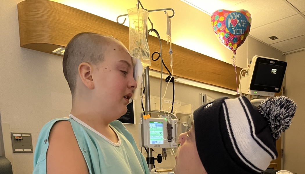 A young hospital patient with a shaved head and eye patch being comforted by a loved one wearing a beanie.