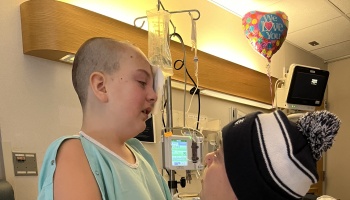 A young hospital patient with a shaved head and eye patch being comforted by a loved one wearing a beanie.