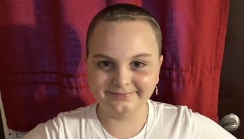 A smiling young person with a shaved head, wearing a white shirt in front of a red curtain.