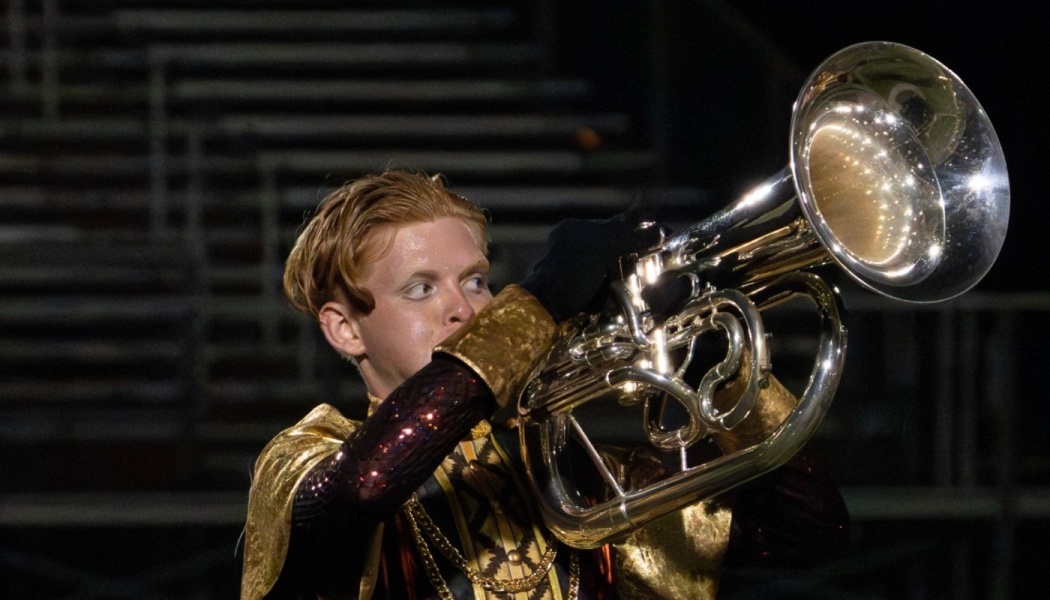 A young performer in an ornate gold marching uniform plays a shiny silver brass instrument at night.