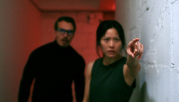 A woman points urgently ahead while a man with glasses looks alarmed in a dimly lit, red hallway.