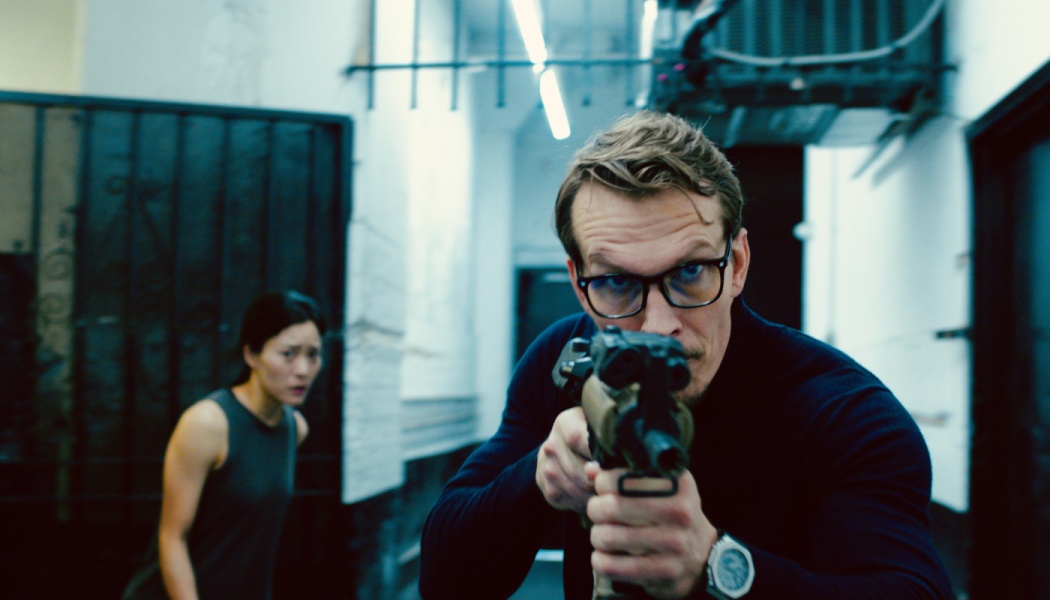A man with glasses aims a gun directly at the camera while an anxious woman watches in a narrow hallway.