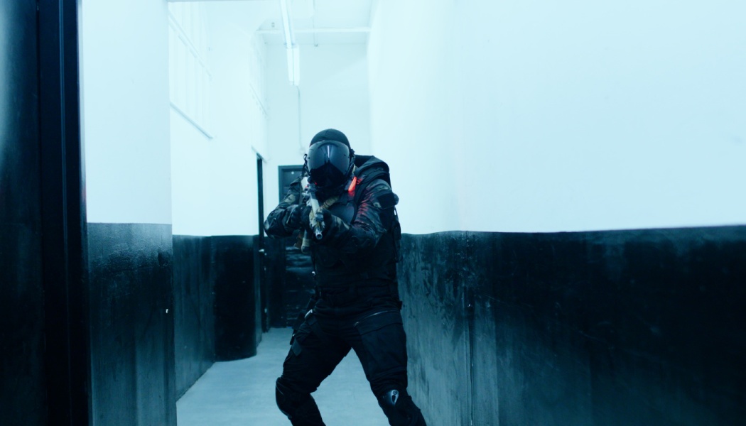 A soldier in black tactical armor and helmet aims a rifle down a narrow, cool-toned hallway.