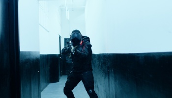 A soldier in black tactical armor and helmet aims a rifle down a narrow, cool-toned hallway.
