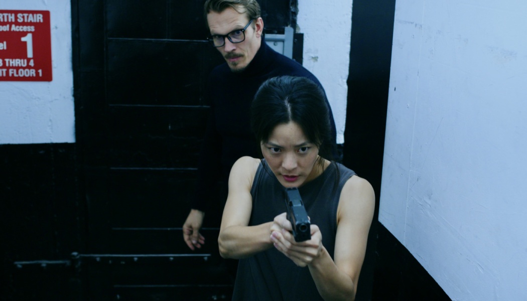 A focused woman aims a handgun forward as a man with glasses stands behind her in a dark stairwell.