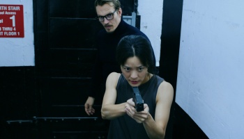 A focused woman aims a handgun forward as a man with glasses stands behind her in a dark stairwell.