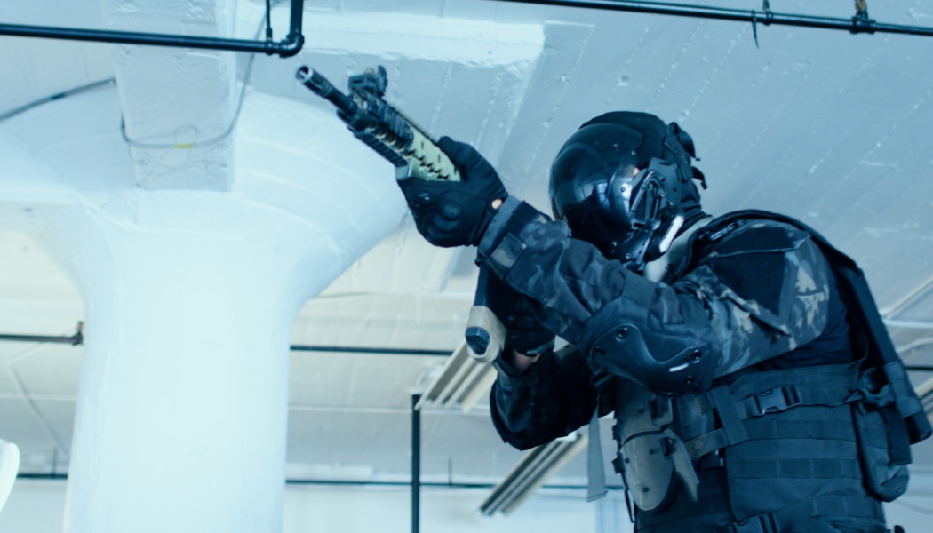 A tactical soldier in dark camouflage gear and a full-face helmet aims a rifle inside a building.
