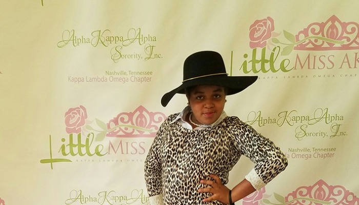 Young girl in a black hat and leopard print top posing in front of a Little Miss AKA sorority backdrop.