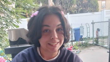 A smiling young person with short dark hair and pink hair bows outdoors in a backyard.