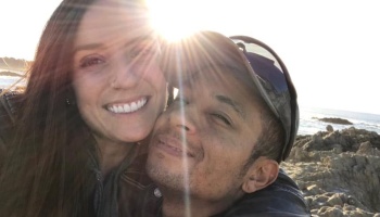 A smiling couple poses together on a rocky coast with bright sunlight flaring behind them.