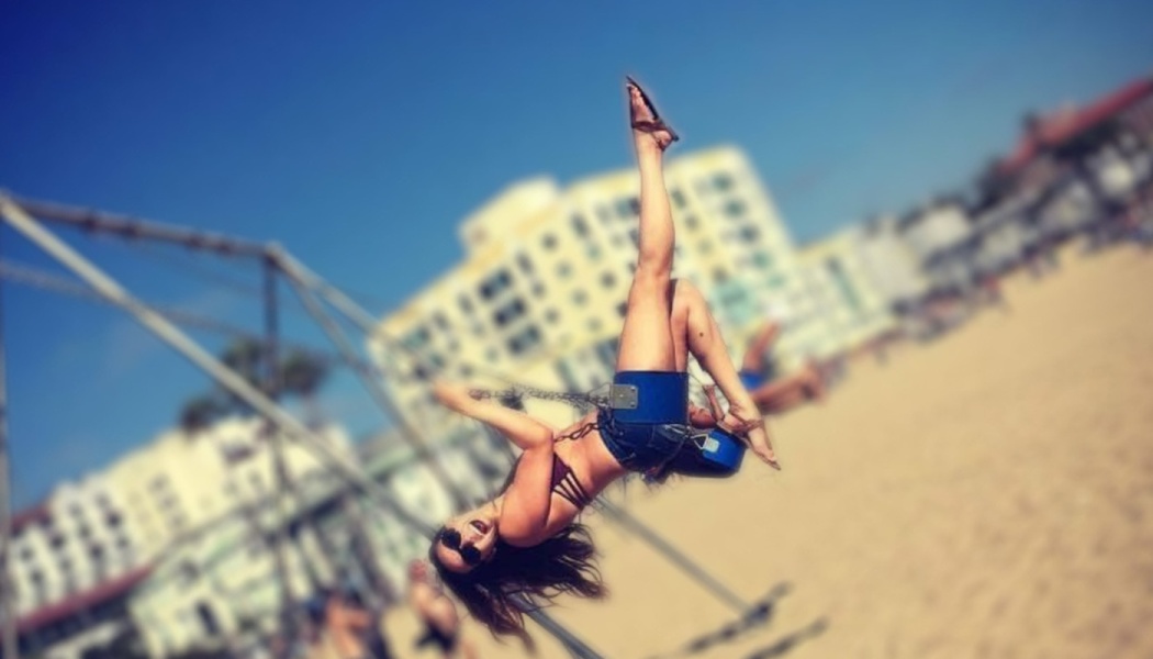 A smiling woman swings playfully on a beach swing set, kicking one leg straight up into the sunny sky.