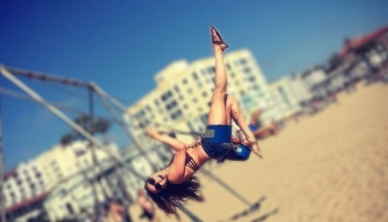 A smiling woman swings playfully on a beach swing set, kicking one leg straight up into the sunny sky.